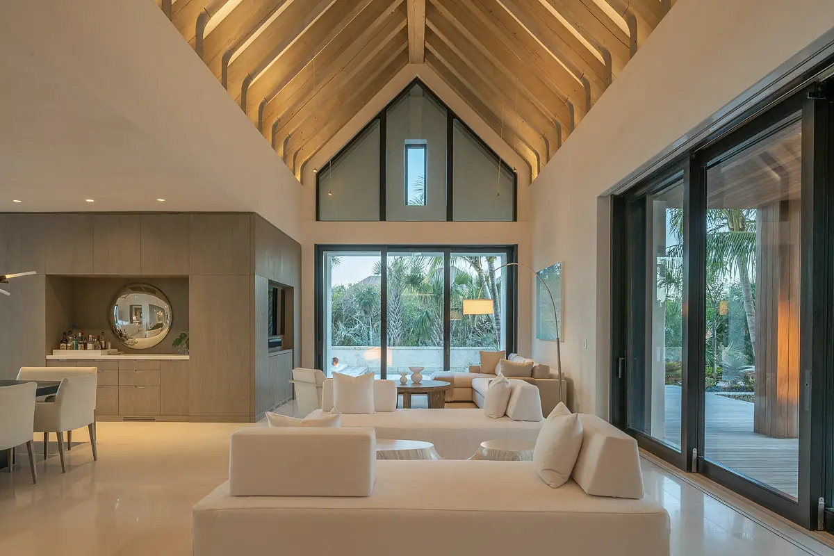 Living room with white sofas, wood coffee table, large windows, high ceiling, and circular mirror