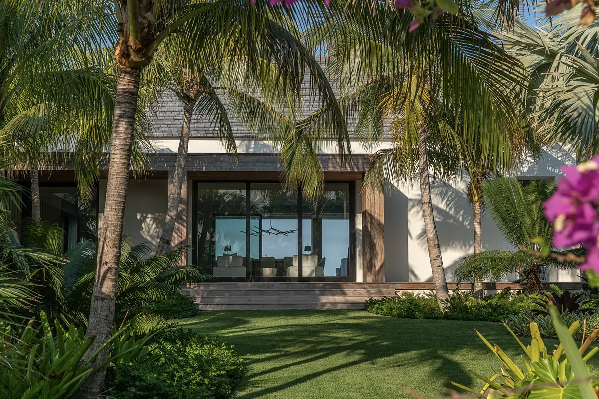 Exterior front of a house surrounded by palm trees and flowering plants with large glass windows and porch.