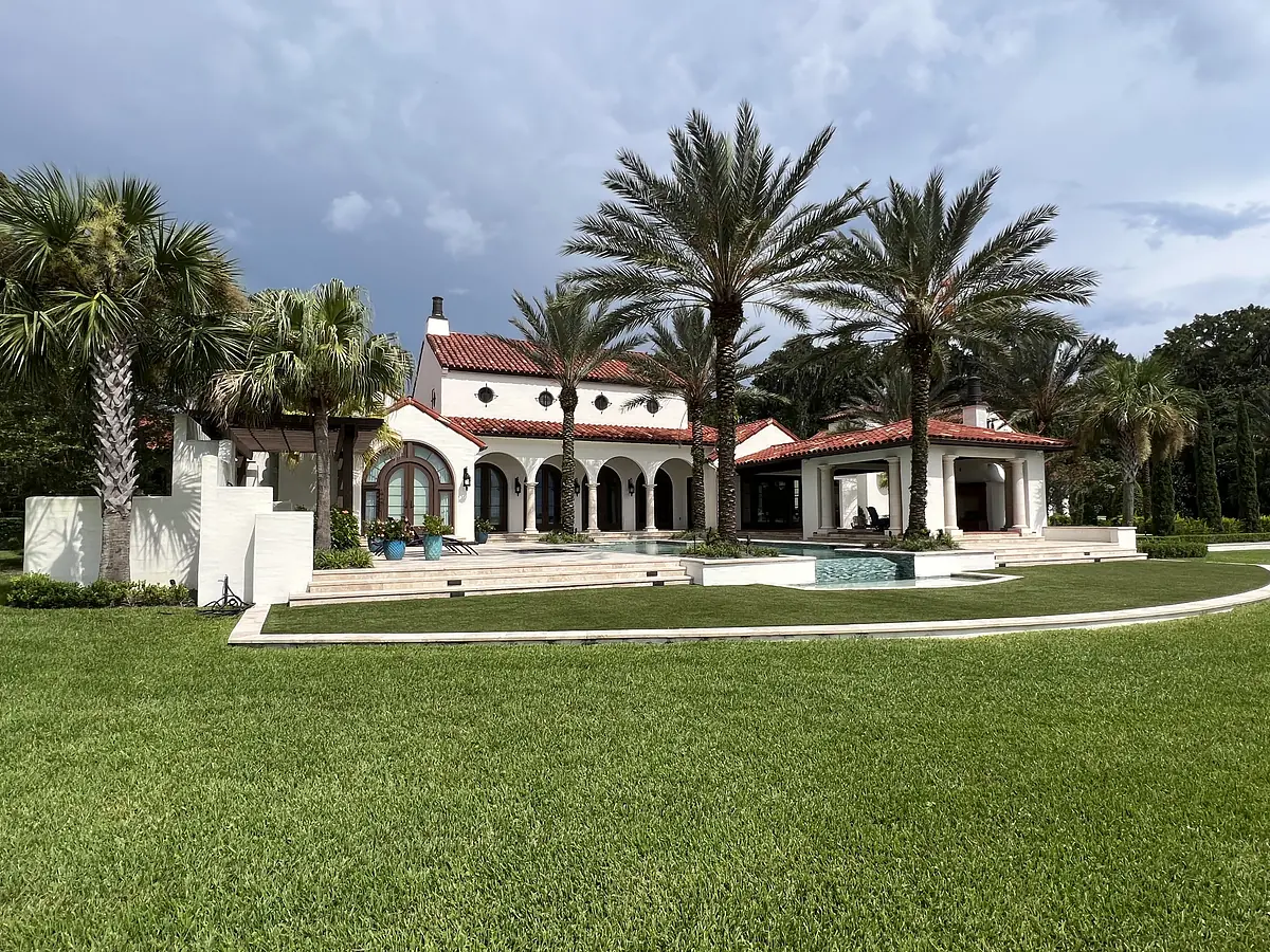 Exterior front view of a house with red-tiled roof, white walls, arched windows, and surrounding palm trees.