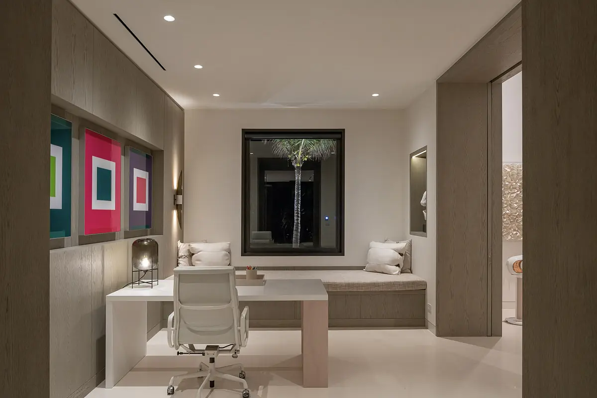 Office with white desk, ergonomic chair, built-in bench, cushions, artwork, and large window.