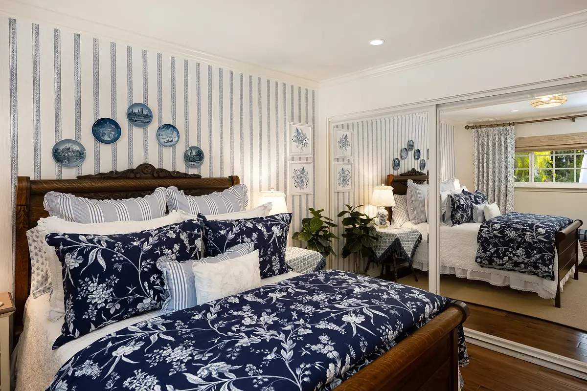 Bedroom with wooden bed, floral navy quilt, nightstands, decorative plates, and window with patterned drapes