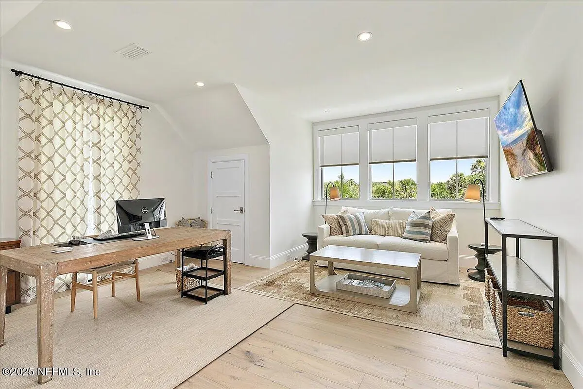 Office with wooden desk, computer, white coffee table, sofa, and large windows with gray shades