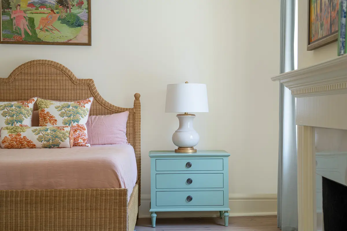 Bedroom with woven bed, pink bedspread, blue nightstand, lamp, and artwork on the walls