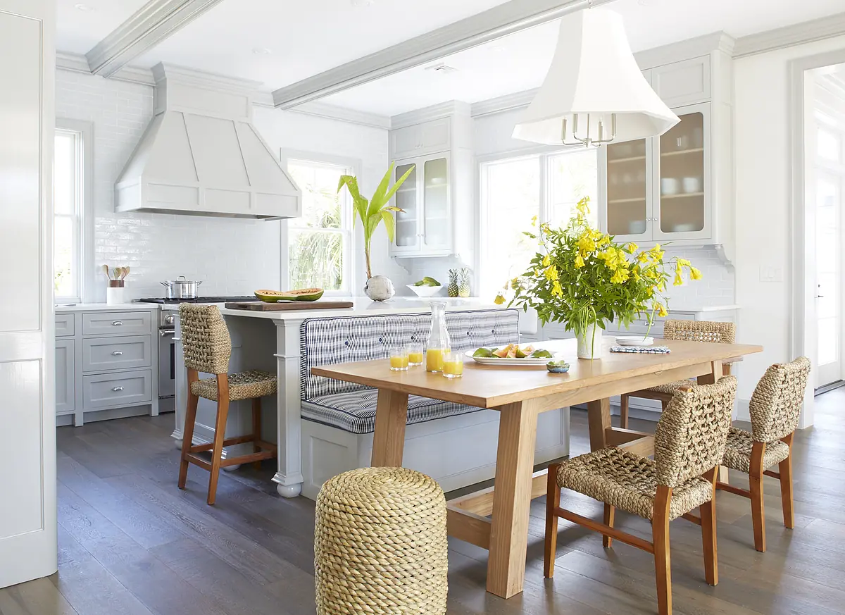 Kitchen with gray island, wooden dining table, woven chairs, and large range hood with flowers and glassware.