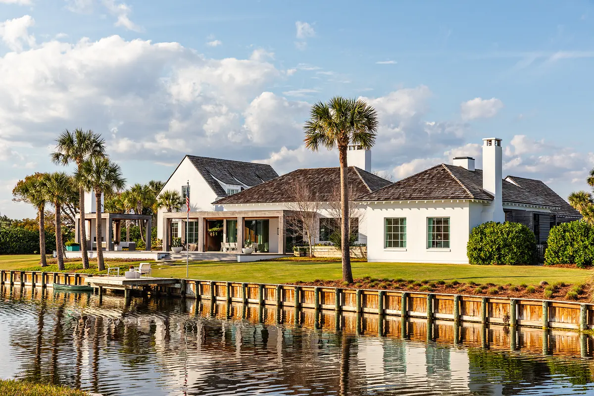 Exterior front view of a white house with pitched roofs, palm trees, and a water body in the foreground.