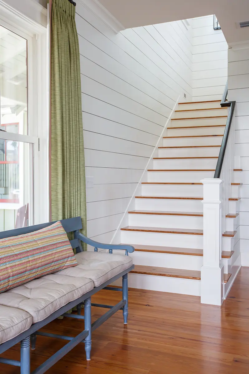 Hallway with blue bench, wooden stairs, white shiplap walls, and green curtains at the window.