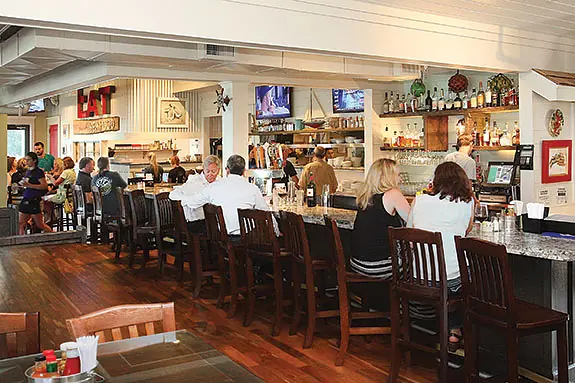 Dining room with long bar counter, wooden stools, tables, decor shelves, and patrons.