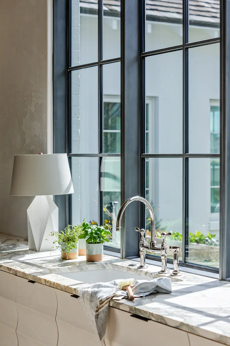 Kitchen with large window, stainless steel sink, marble countertop, potted plants, and geometric lamp.