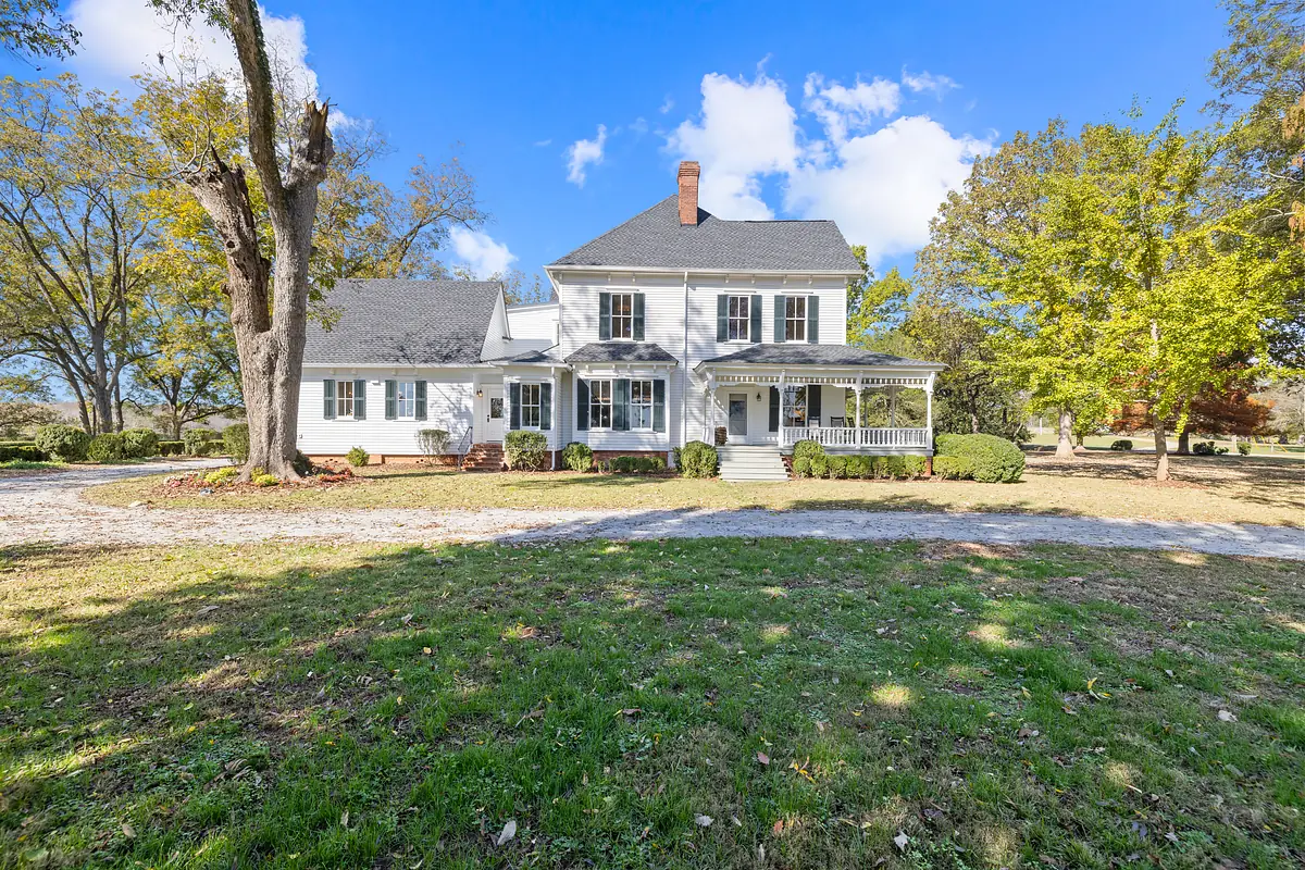Front exterior of a two-story white house with gray roof, porch, driveway, shrubs, and trees.
