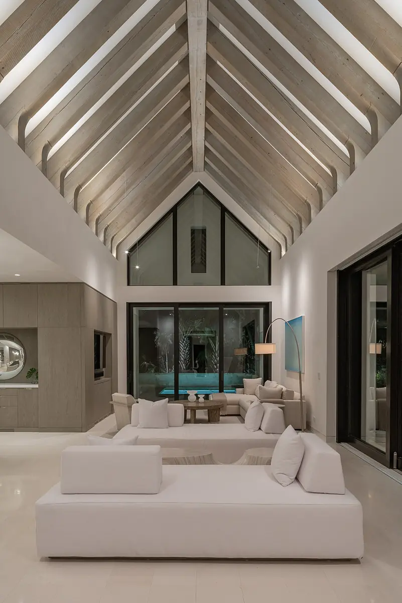 Living room with white sofas, wooden coffee table, large windows, and peaked ceiling with wooden beams overlooking a pool.