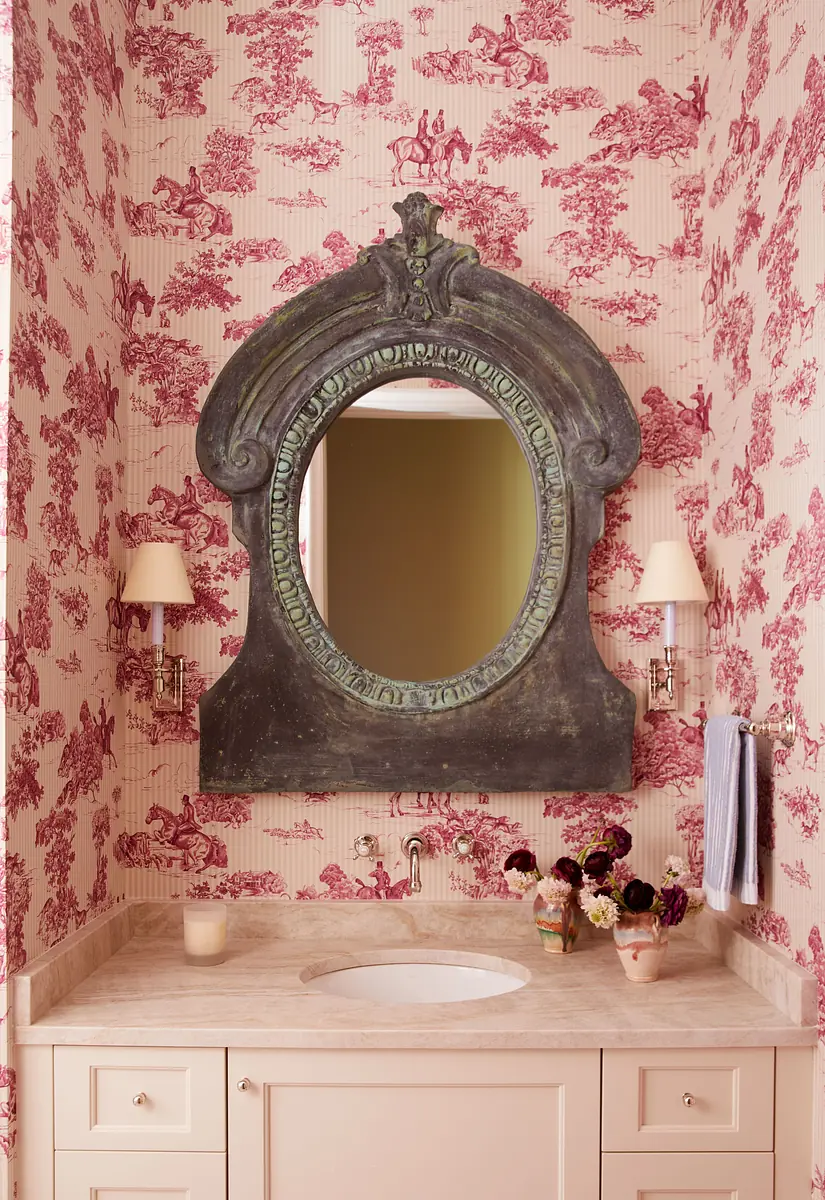 Bathroom with ornate mirror, floral wallpaper, stone countertop, wall sconces, and a vase of flowers