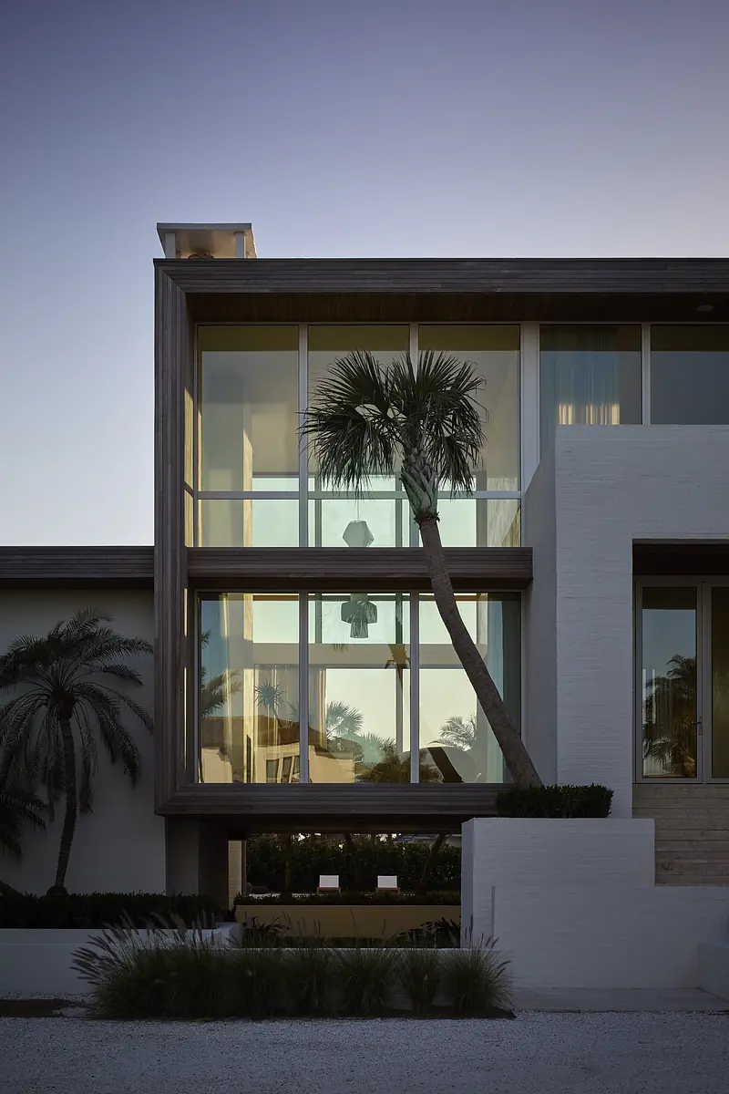 Exterior view of modern building with large glass windows, wooden frame, white facade, and palm tree.