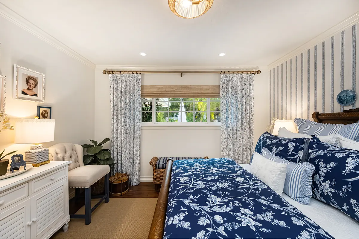 Bedroom with blue floral bedding, white dresser, armchair, area rug, curtains, and artwork on walls.