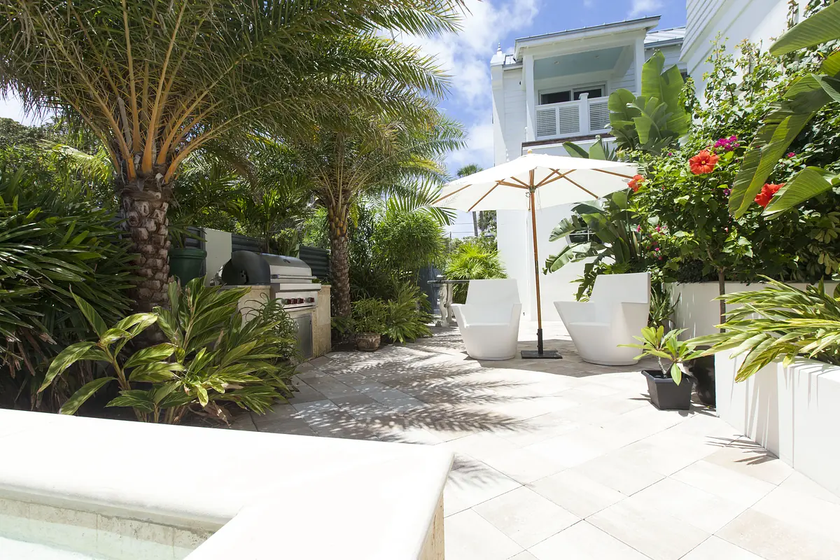 Garden with white chairs, umbrella, stone patio, tropical plants, and a grill against a wall.