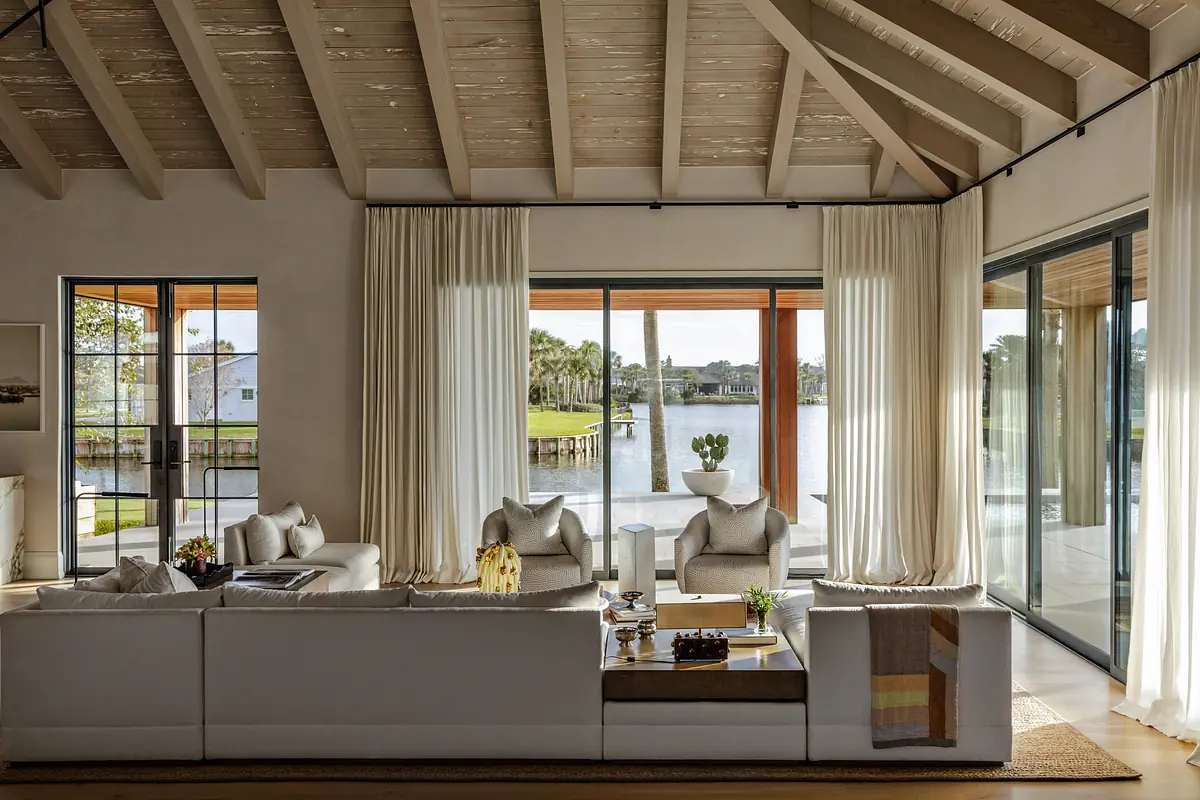 Living room with sectional sofa, coffee table, large windows, and exposed beam ceiling overlooking water.