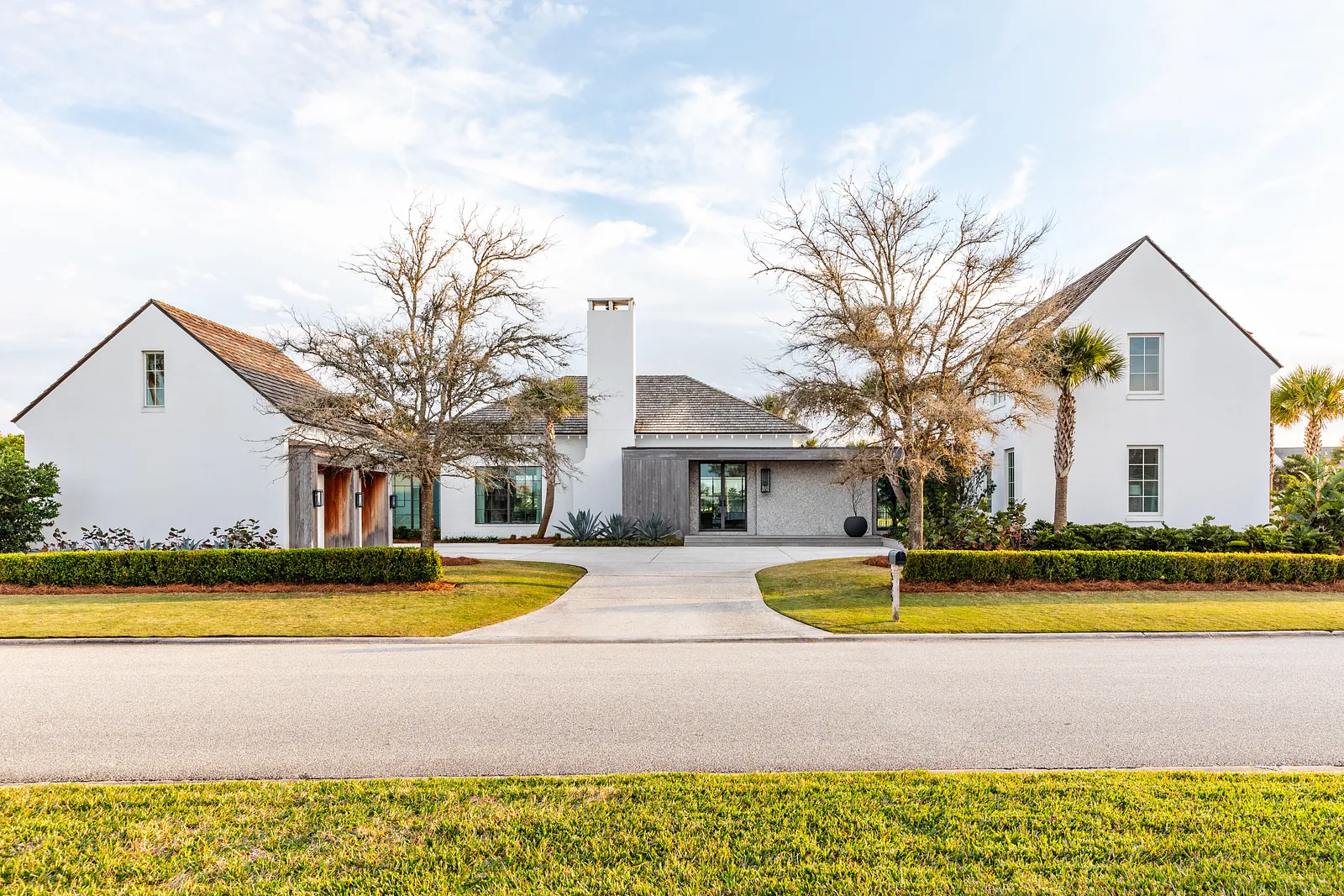 Front exterior of a house with gable roofs, central chimney, driveway, palm trees, and landscaped greenery.