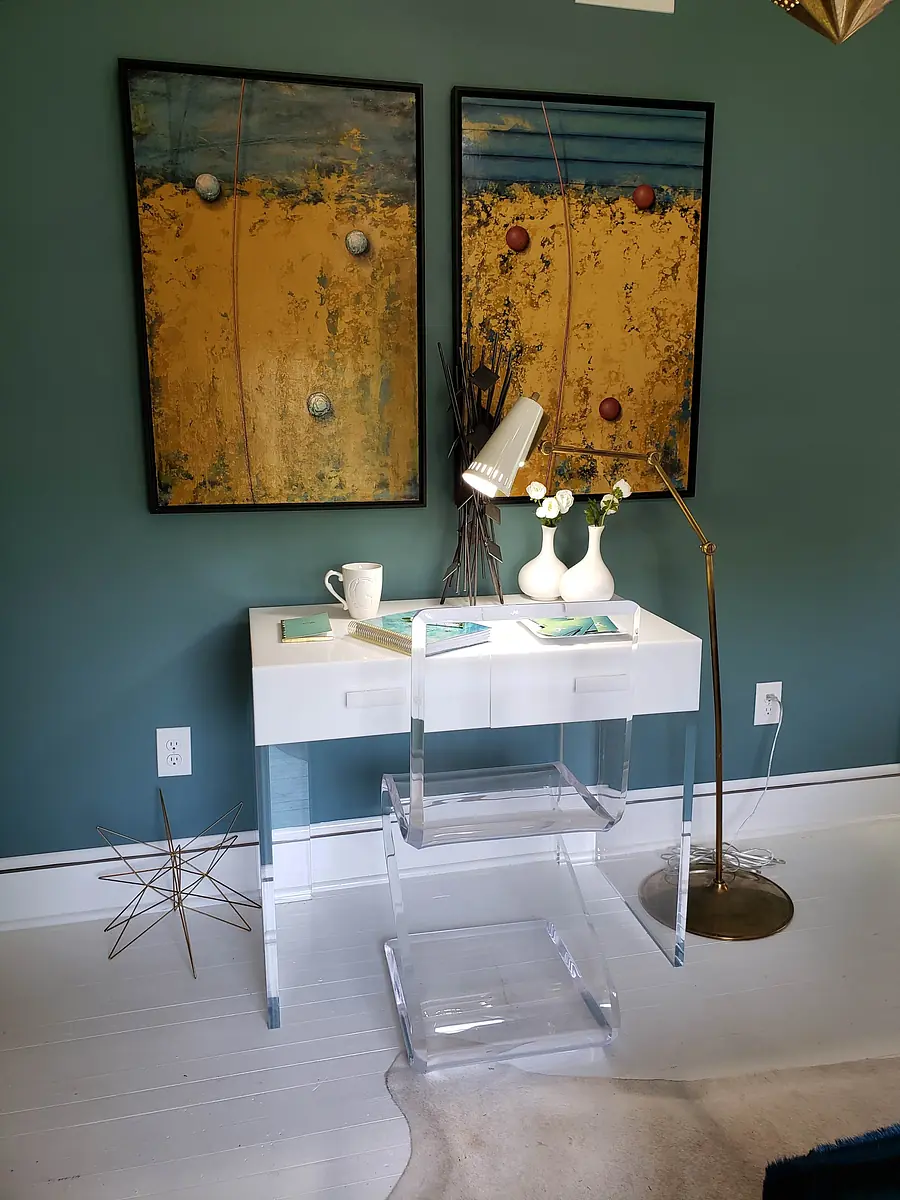 Office with white desk, two paintings, floor lamp, flowers, and acrylic shelf against a teal wall.