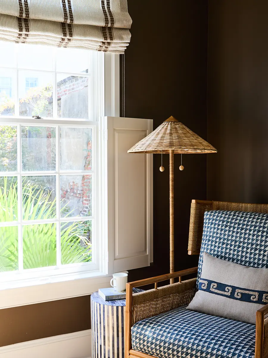 Corner setting with blue and white chair, side table, straw lamp, and window with greenery view