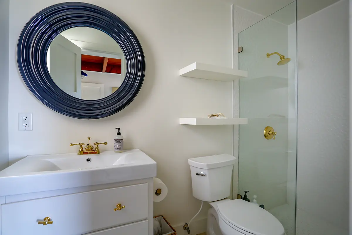 Bathroom with white vanity, gold faucet, clear glass shower, and round mirror above the sink.