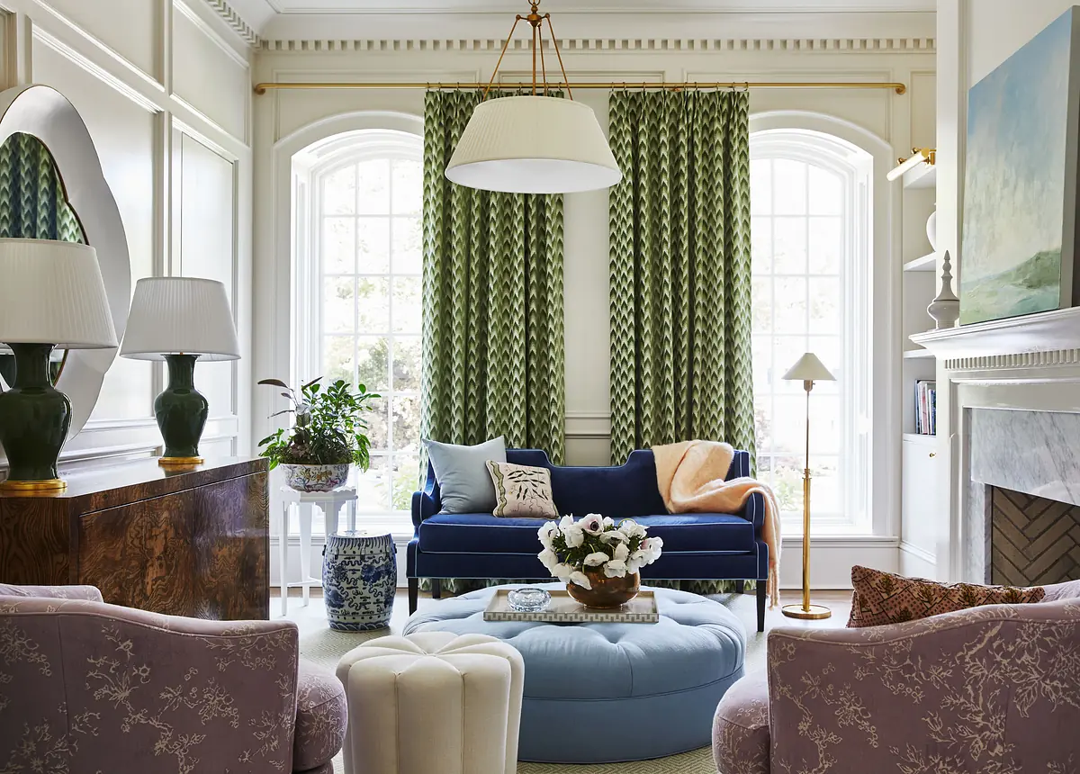 Living room with blue sofa, purple chairs, round ottoman, coffee table, plants, and large windows with curtains.