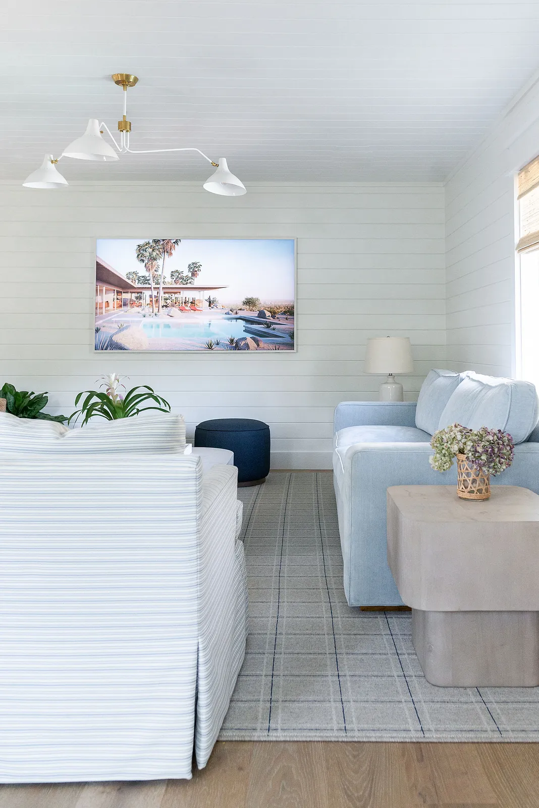 Living room with white sectional, light blue sofa, plaid rug, blue ottoman, artwork, and potted plant.