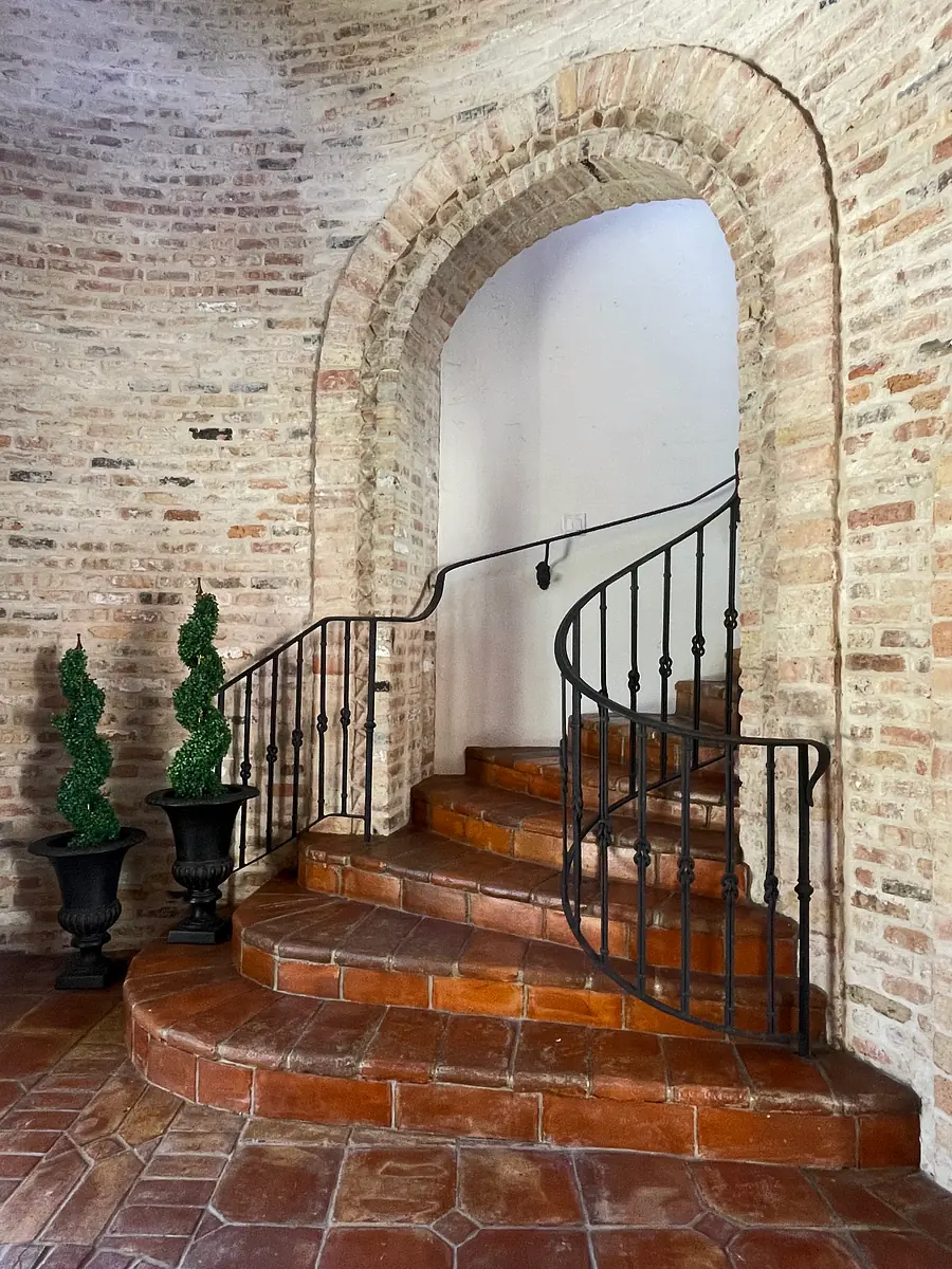 Hallway with curved brick walls, terracotta floor, wrought-iron railing, and topiary planters.