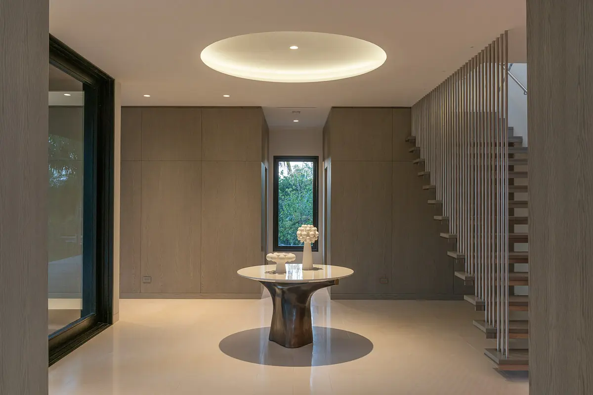 Foyer with round table, vase, staircase, large windows, and light wood paneling.