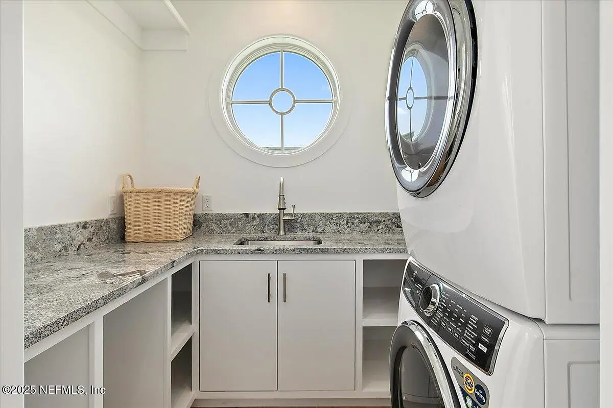 Laundry room with stacked washer and dryer, granite countertop, sink, and white cabinetry with circular window.
