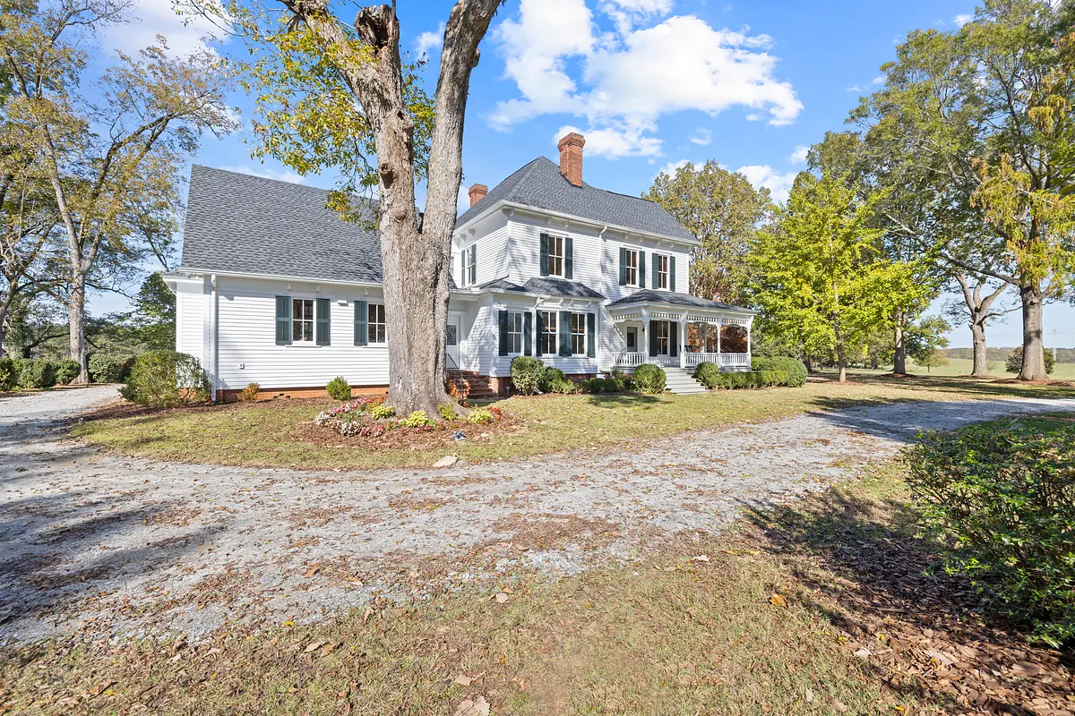 Front view of a two-story house with white siding, black roof, wraparound porch, and gravel driveway.