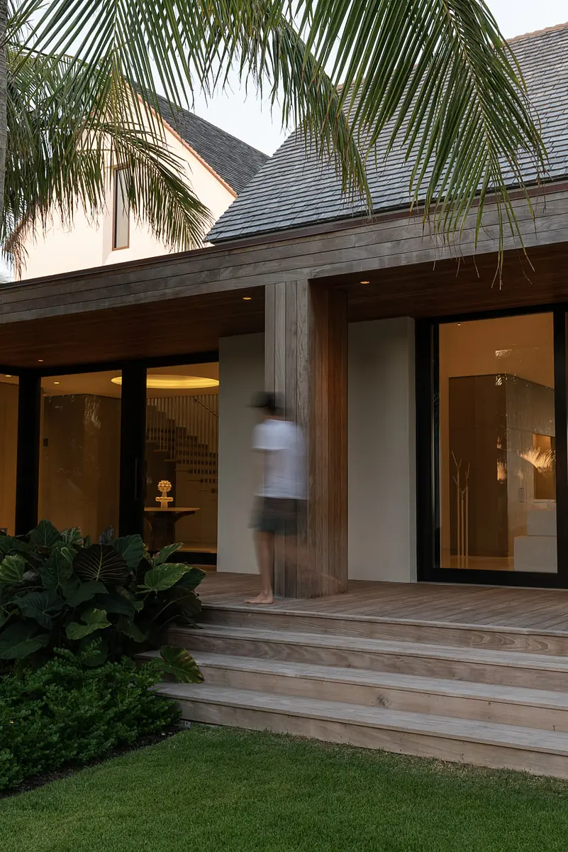 Exterior front area with wooden deck, glass doors, tropical foliage, and gabled roof.
