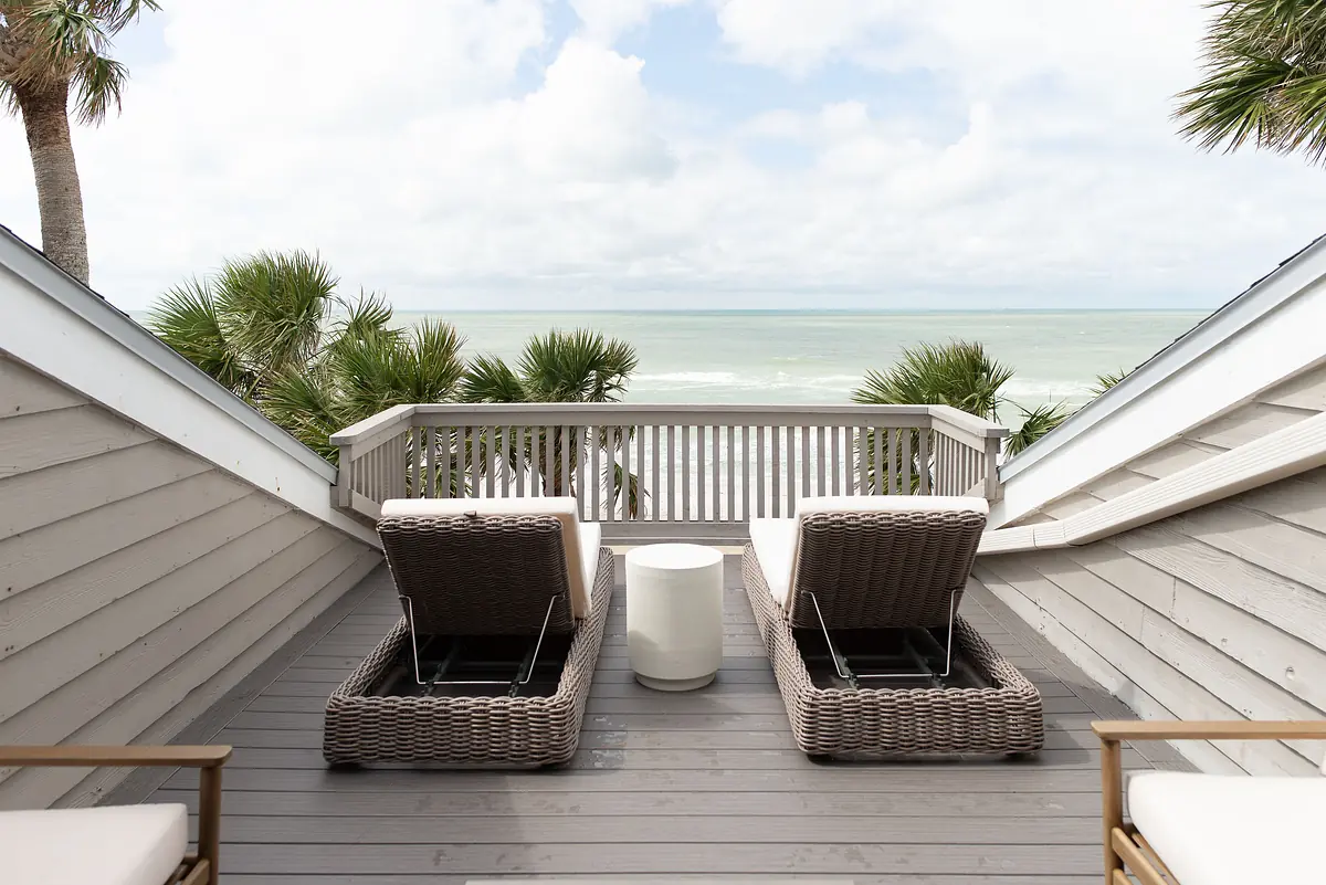 Deck with two wicker lounge chairs, a white side table, and ocean view surrounded by palm trees.