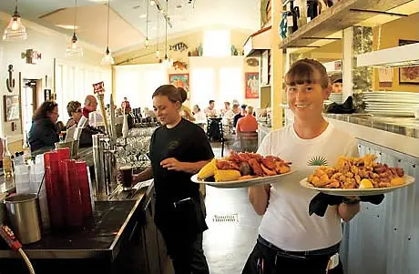 Busy restaurant interior with bar counter, tables, chairs, staff serving food, and decorative wall art.