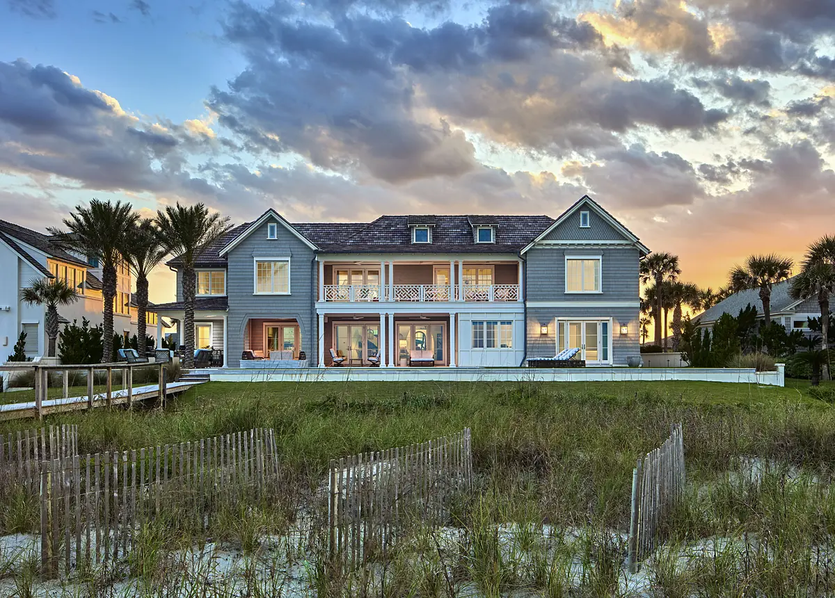 Rear exterior view of a two-story gray house with balcony, surrounded by grass and palm trees near a beach.