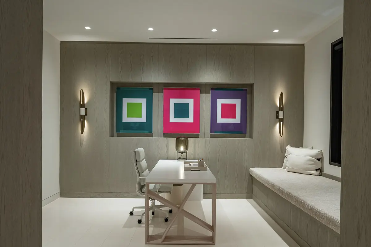 Office with white desk, ergonomic chair, built-in bench, colorful wall art, and light wood paneling