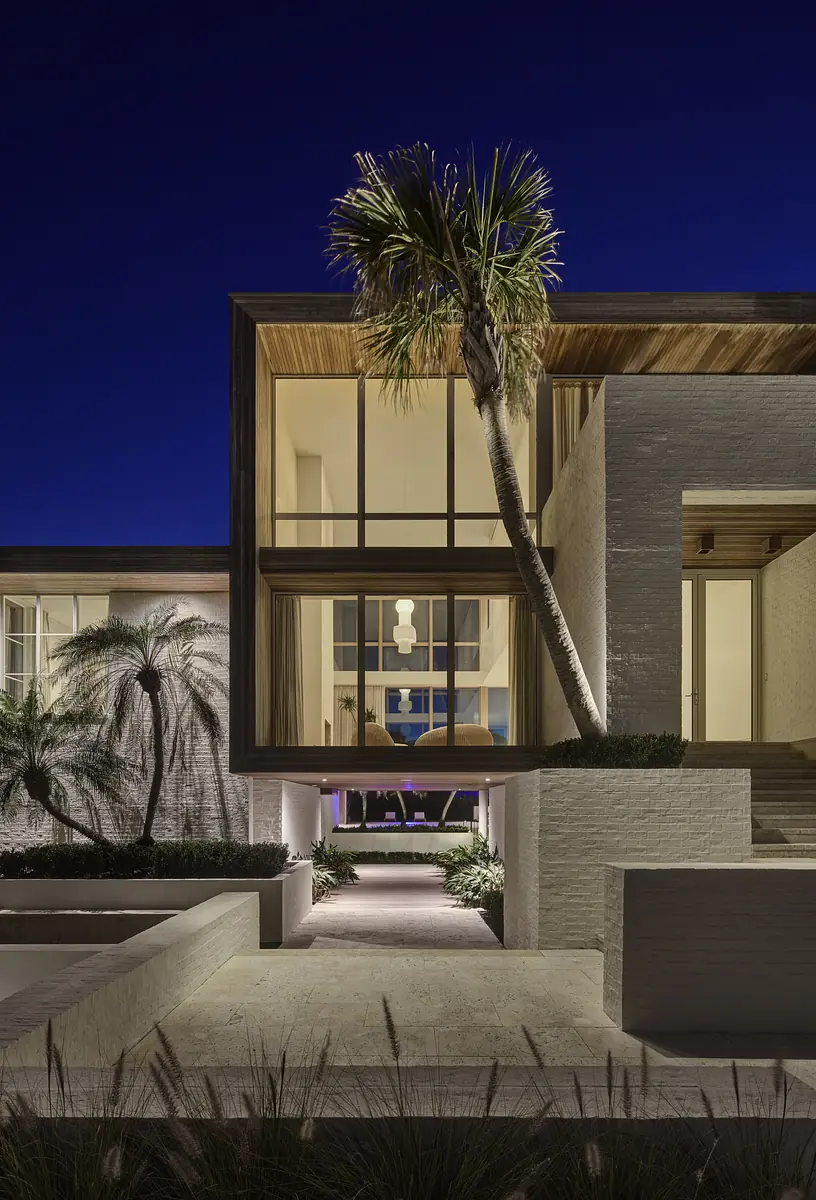 Front exterior of a modern house with large windows, wooden overhang, palm trees, and path leading to the entrance.