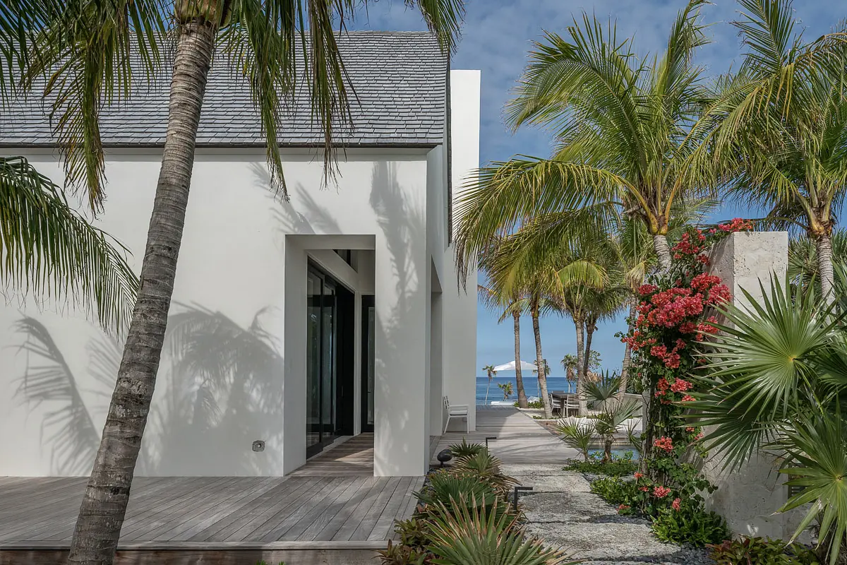 Exterior front view of a modern house with palm trees, flowers, and a stone pathway to the ocean.