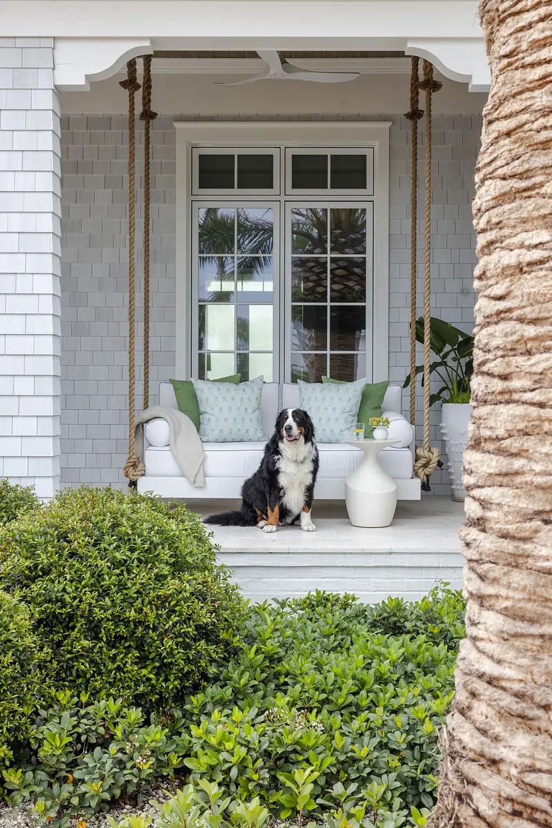Patio with white swing sofa, green cushions, side table, potted plants, and surrounding shrubs.