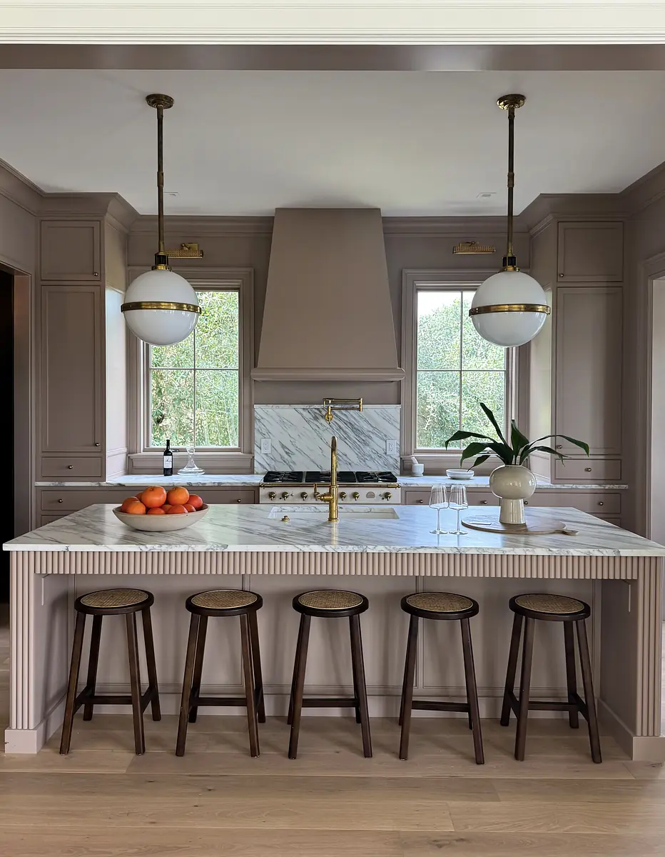 Kitchen with marble island, wooden stools, gray cabinetry, two pendant lights, and large windows.