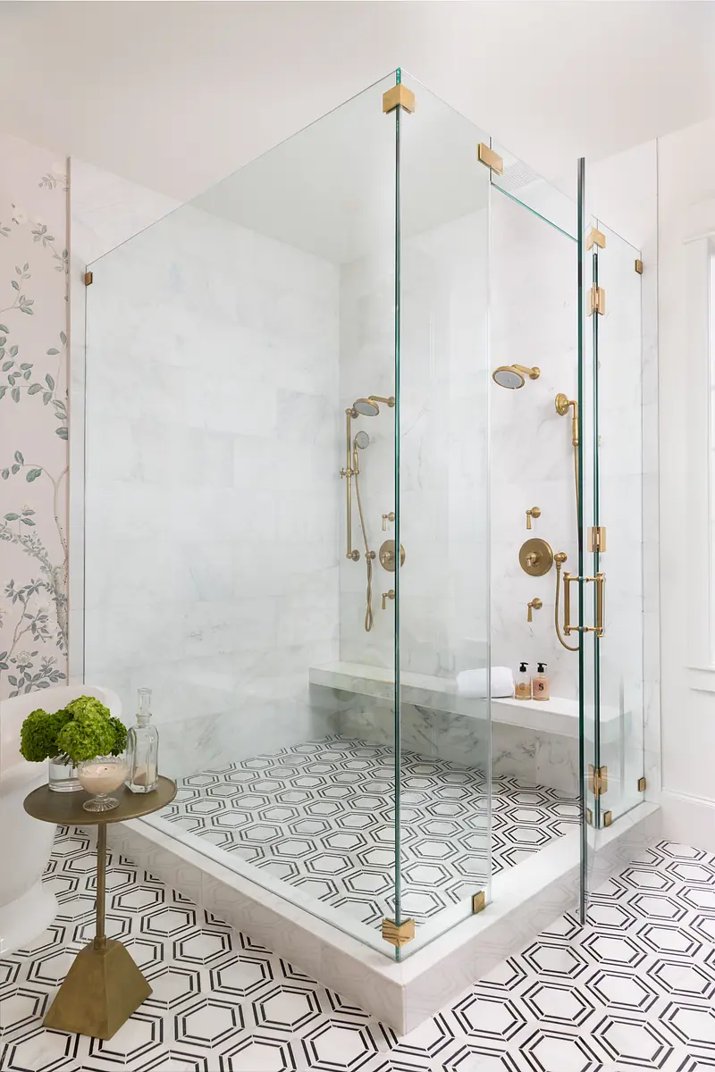 Bathroom with glass shower, brass fixtures, marble walls, and geometric black and white tile floor.