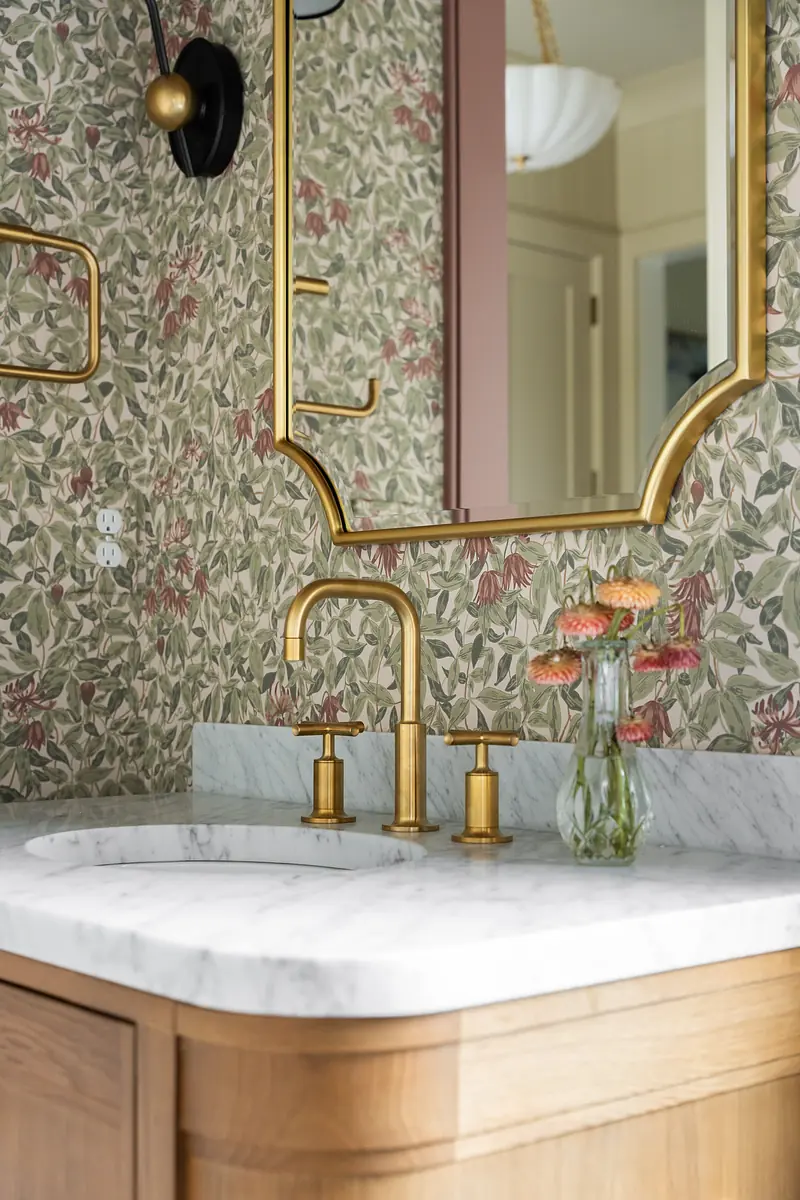 Bathroom with marble countertop, gold faucet, mirror, and floral wallpaper.