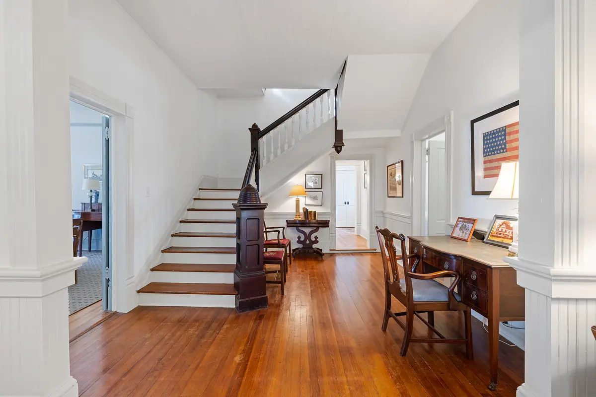 Hallway with wooden staircase, hardwood floor, desk, chair, and wall-mounted art including an American flag.