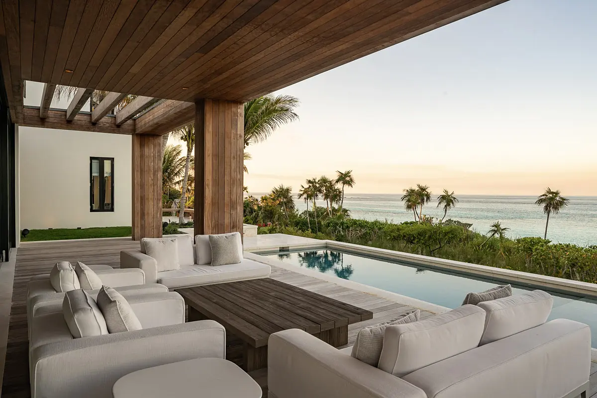 Patio with sectional sofa, coffee table, swimming pool, and lush greenery under wooden overhang.