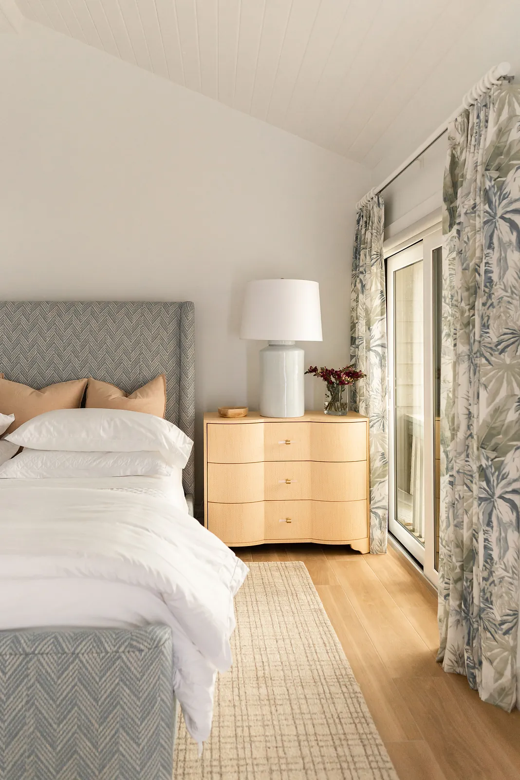 Bedroom with patterned headboard, white bedding, nightstand, blue lamp, and floral drapes.
