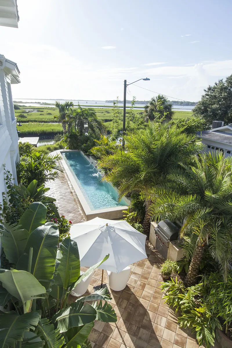 Patio with pool, palm trees, white chairs, umbrella, beige tiles, and barbecue grill with water view.