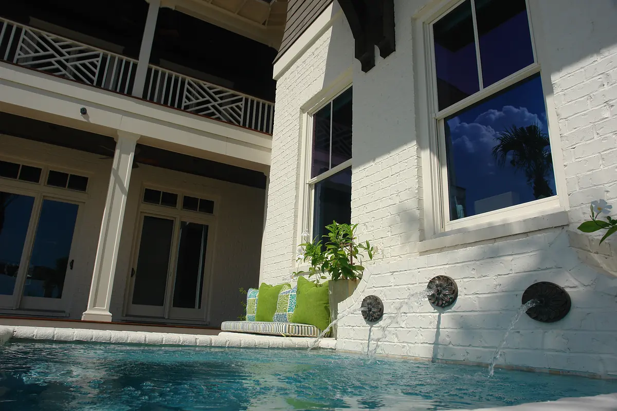 Patio with swimming pool, water fountains, green pillows, and white brick wall with wooden beams.