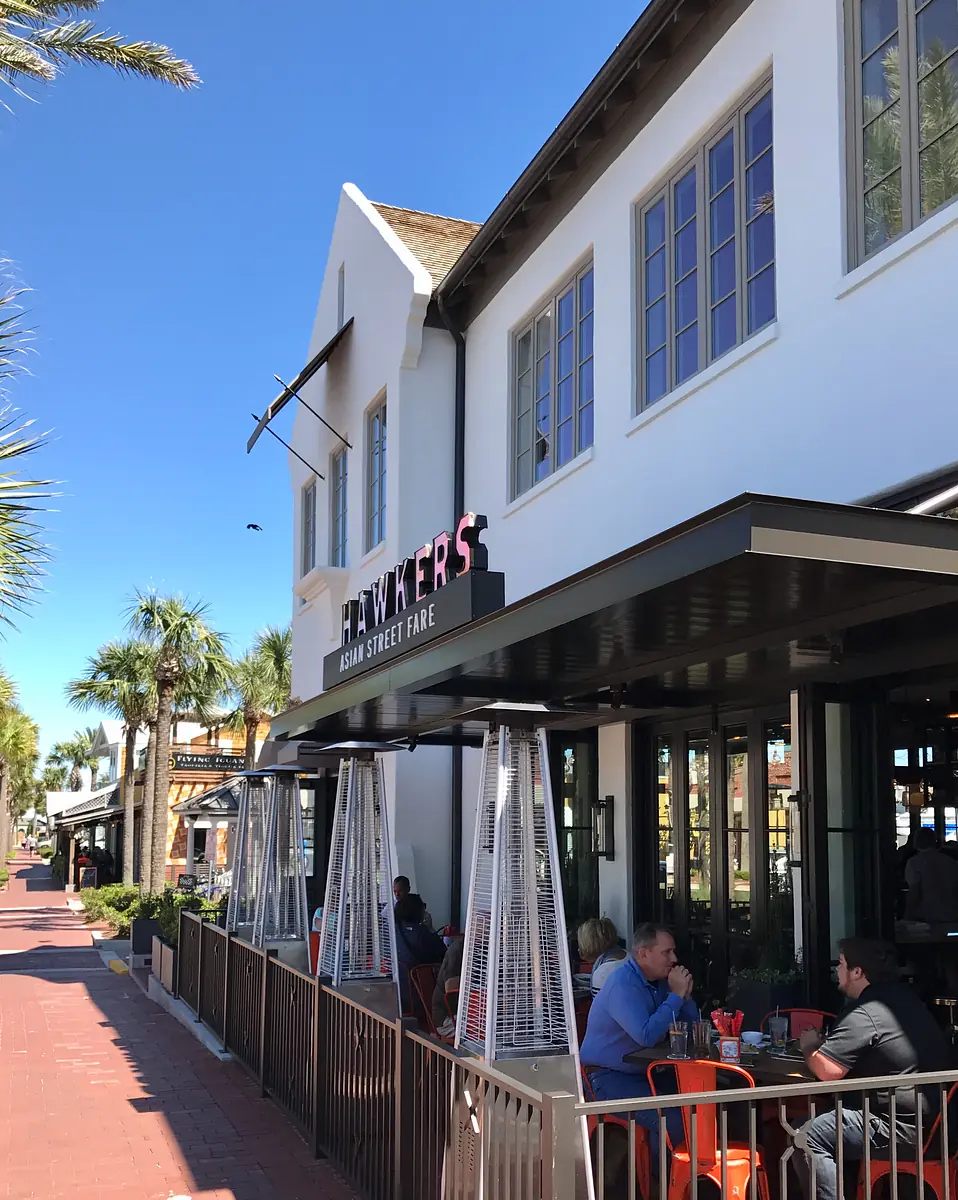 Exterior front view of 'Hawkers Asia Street Fare' with seating area, palm trees, and large windows.