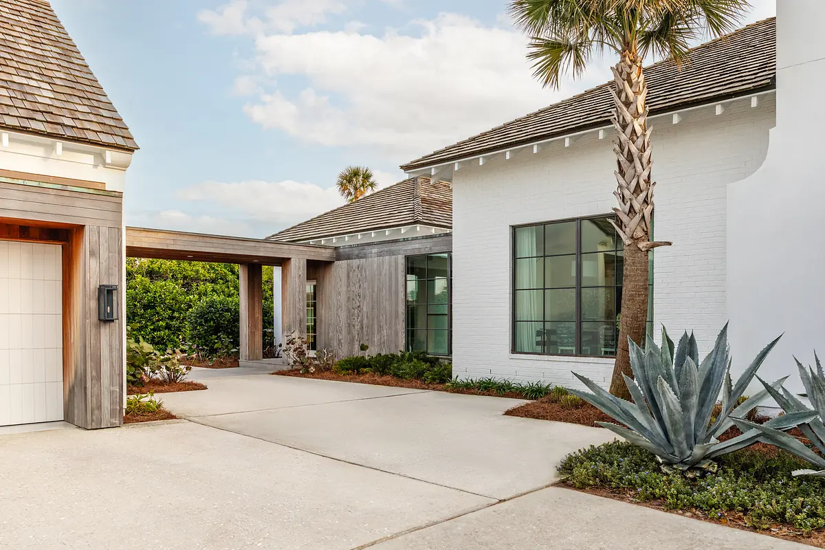 Exterior front view of a home with paved driveway, palm trees, agave, and a combination of white brick and wood siding.