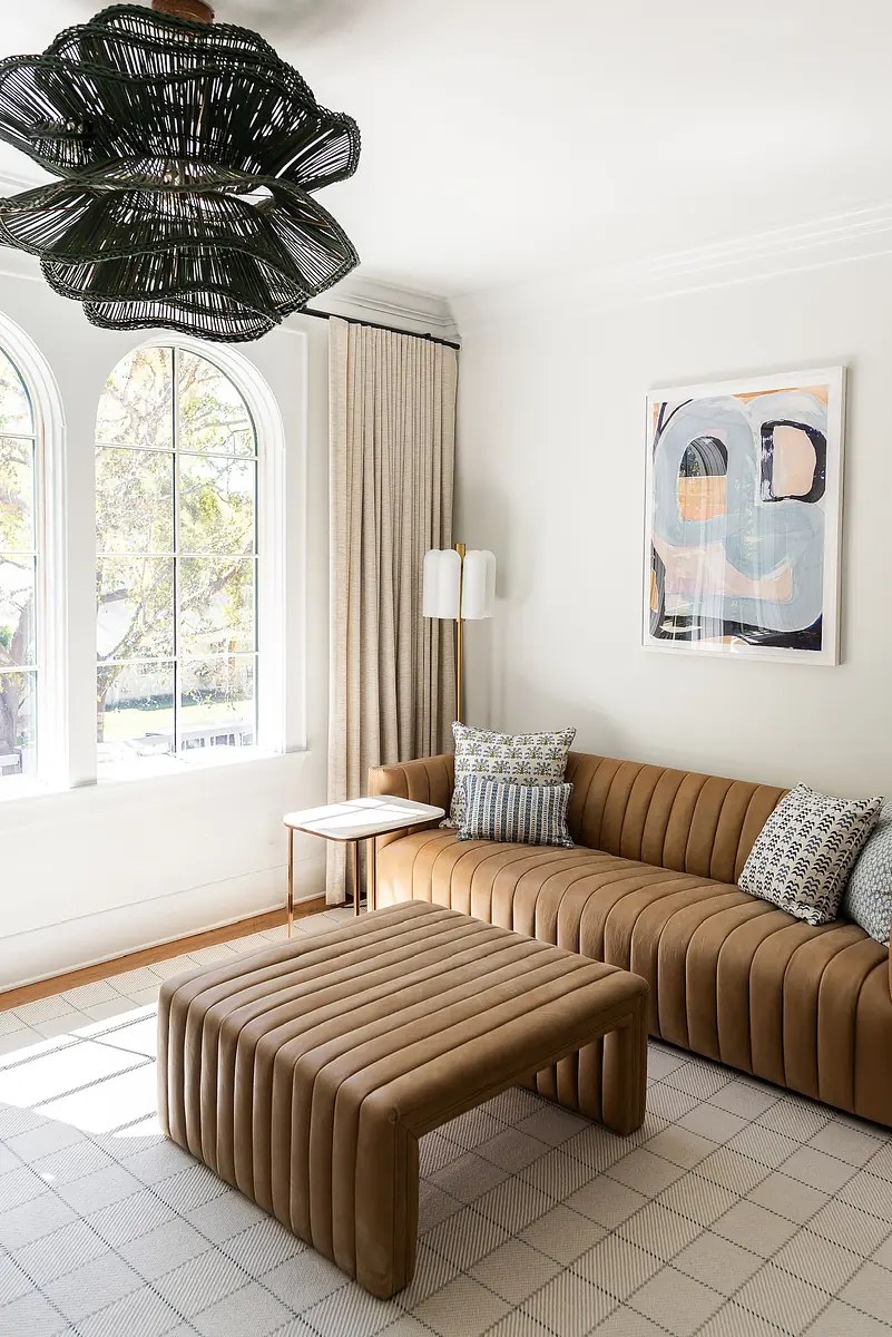 Living room with brown curved sofa, striped coffee table, large windows, artwork, and side table with lamp.