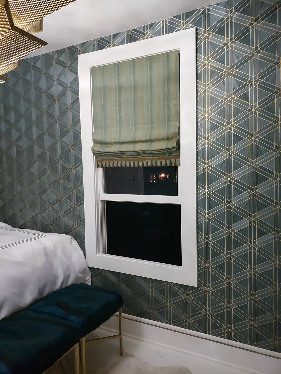 Bedroom corner with window, green roman shade, geometric wallpaper, and cushioned bench in front.