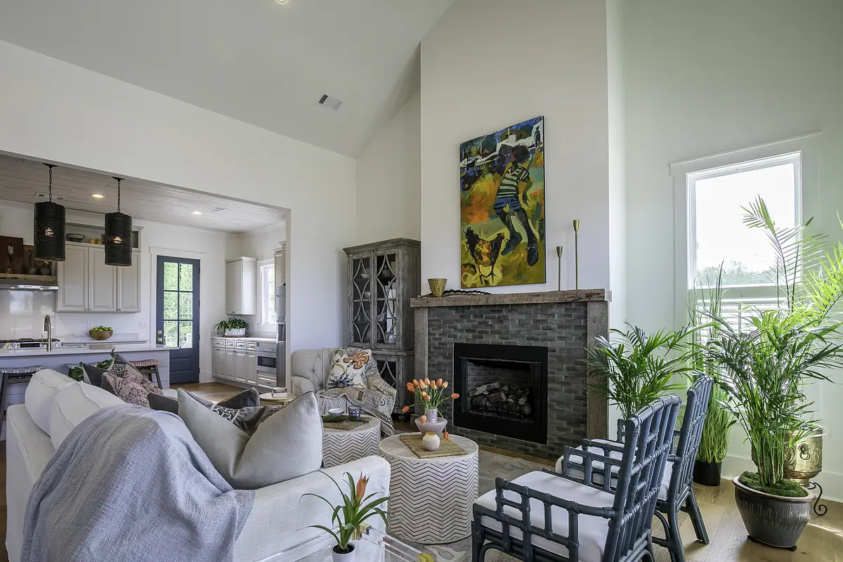 Living room with white sofa, blue chairs, coffee table, stone fireplace, large painting, and kitchen area in background.